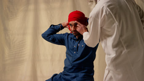 Studio-Shot-Of-Senior-And-Younger-Sikh-Men-Tying-Fabric-For-Turban-Against-Plain-Background